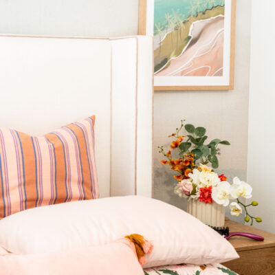 A neatly made bed with the COCO Bedhead, pink and striped pillows, a bedside table featuring a floral arrangement, and a framed coastal artwork on the wall.