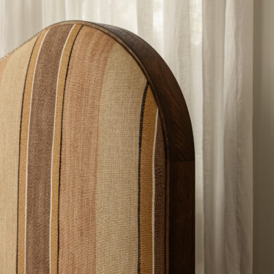 Agnes upholstered headboard features vertical beige and brown stripes, a wooden trim, and is set against sheer white curtains.