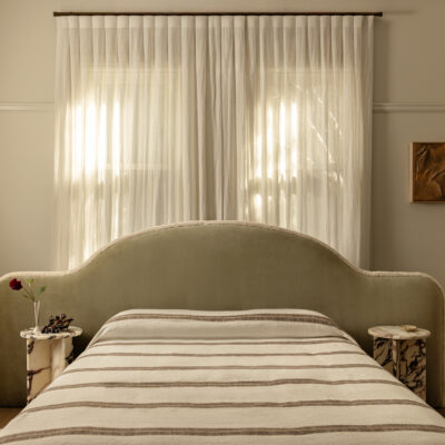 A neatly made bed featuring the Paloma striped blanket, paired with a velvet headboard, two compact bedside tables, and sheer curtains draping over a sunlit window.