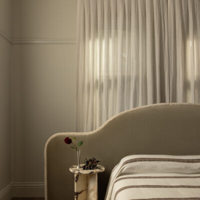 Paloma minimalist bedroom featuring a curved beige Paloma headboard, striped bedding, marble bedside table, single flower in a vase, and sheer curtains that filter in soft daylight.