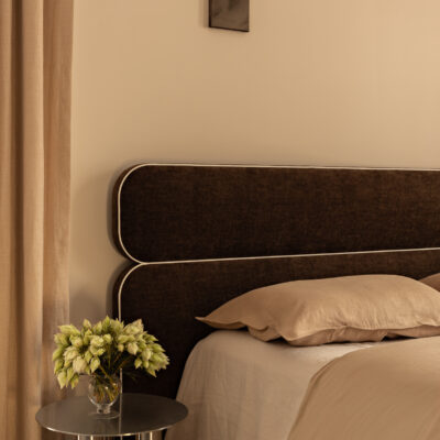 Minimalist bedroom featuring the Jean upholstered headboard in a dark hue, beige walls, light bedding, a small round metal bedside table, and a vase of green flowers.