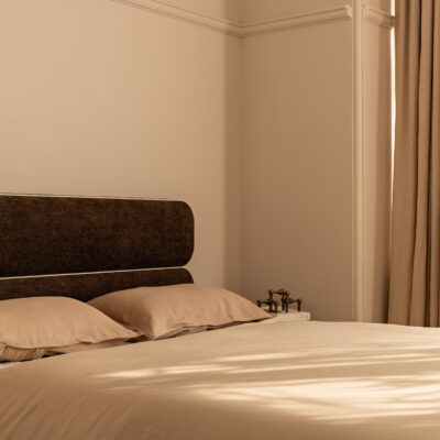 A neatly made bed featuring the Jean beige sheets and pillows sits in a softly lit room with beige walls, a brown headboard, and closed beige curtains.