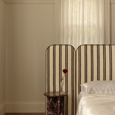 The Willow striped upholstered headboard stands behind a neatly made bed, accompanied by a small marble bedside table holding a single red rose in a glass vase; sheer curtains softly veil the window behind.