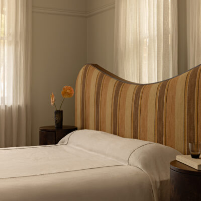 Agnes bed with a striped, curved headboard, paired with two round wooden bedside tables—one holding a vase of flowers, the other set with a book and glass—all bathed in soft light filtered through sheer curtains covering the windows.