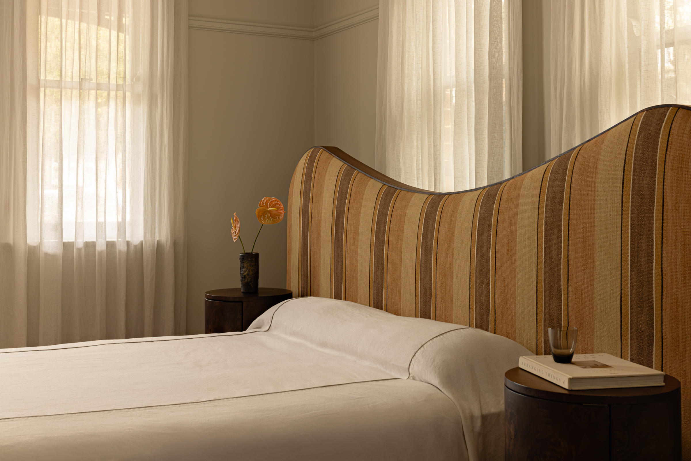 Agnes bed with a striped, curved headboard, paired with two round wooden bedside tables—one holding a vase of flowers, the other set with a book and glass—all bathed in soft light filtered through sheer curtains covering the windows.