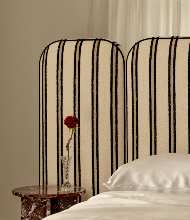 A single red rose in a glass vase sits on a round marble bedside table next to a bed with a white pillow and a striped headboard.