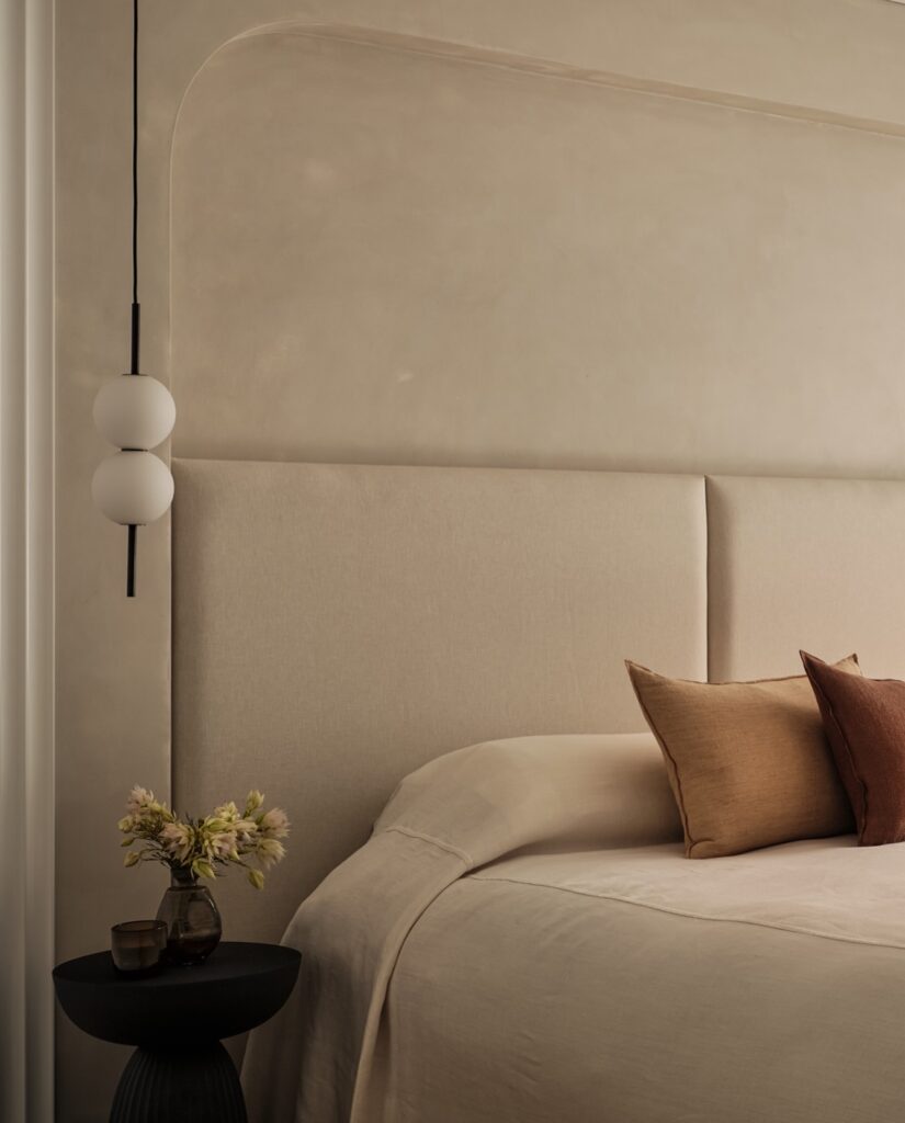 Minimalist bedroom with beige linen bedding, upholstered headboard in Bedhead fabrics, two brown accent cushions, a small black bedside table with a vase of flowers, and a modern hanging light fitting with two round bulbs—a serene space inspired by elegant Australian homes.